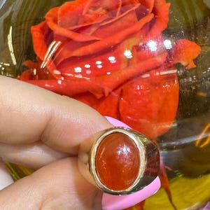 May include: A gold ring with an oval, orange gemstone. The ring has a simple, classic design with a polished gold band. The gemstone is a vibrant orange color and is set in a bezel setting. The ring is shown against a background of a red rose.