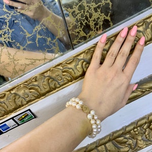May include: A double strand pearl bracelet with a gold charm. The bracelet is on a person's wrist. The person's nails are painted a light pink color.
