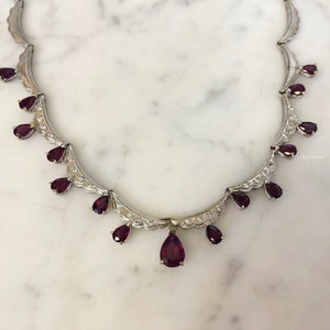May include: A silver necklace with a pear-shaped ruby pendant and smaller ruby accents. The necklace is set with small diamonds and has a delicate design.
