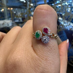 May include: Gold ring with three gemstones: a round emerald, a pear-shaped ruby, and a round diamond. The ring is set with smaller diamonds around each gemstone.