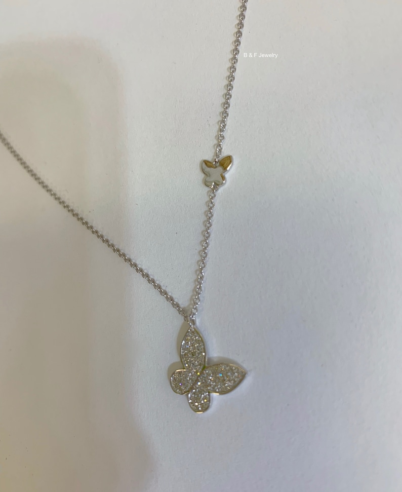 May include: A silver chain necklace with a small butterfly charm at the top and a larger butterfly charm with clear stones at the bottom.  B & F Jewelry is printed on the image.