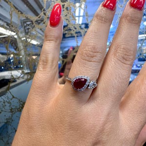 May include: A silver ring featuring a pear-shaped, deep red gemstone surrounded by a halo of smaller, clear stones. The ring also has two smaller, rectangular clear stones on either side of the main stone. The ring is displayed on a hand with red nail polish.