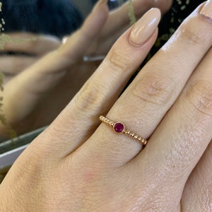 May include: A gold-colored ring with a small, round, red gemstone set in the center. The band features a beaded design. The ring is shown on a person's finger.