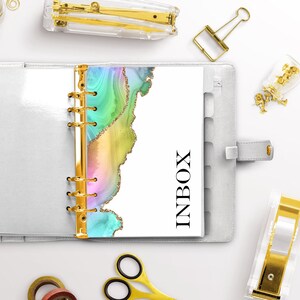 May include: A white planner with a gold ring binder and a colorful abstract design on the cover. The design features a rainbow of colors with gold glitter accents. The word "INBOX" is printed in black on the cover.