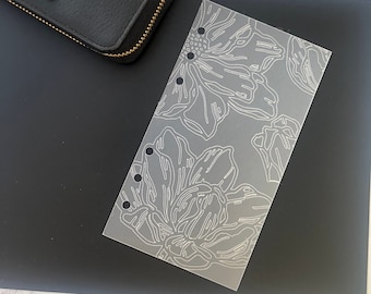 May include: A clear plastic planner insert with a floral design. The design features white outlines of flowers and leaves on a translucent background.