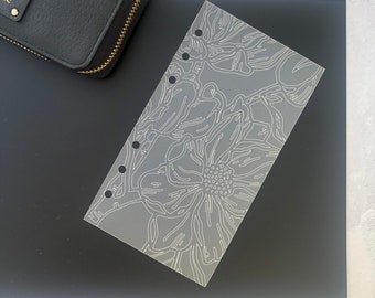May include: A clear plastic planner stencil with a floral design. The stencil has six holes on the left side for a planner.