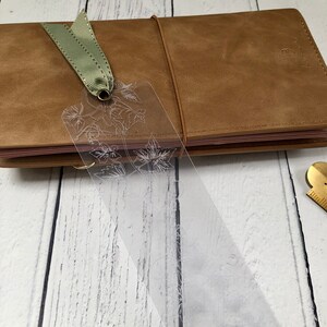 Könnte beinhalten: Ein durchsichtiges Plastik-Lesezeichen mit einem Blumenmuster. Das Lesezeichen ist an einem braunen Leder-Notizbuch mit einem grünen Band befestigt.