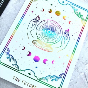 May include: A white card with a rainbow border and gold lettering. The card features a stylized illustration of a crystal ball with an eye inside, surrounded by stars and crescent moons. The text "THE FUTURE" is printed in gold at the bottom of the card.