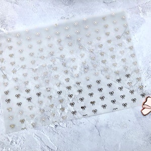 Może przedstawiać: Arkusz przezroczystego papieru samoprzylepnego z powtarzającym się wzorem małych złotych kokardek. Większa różowa kokardka ze złotymi akcentami znajduje się w prawym dolnym rogu obrazu.