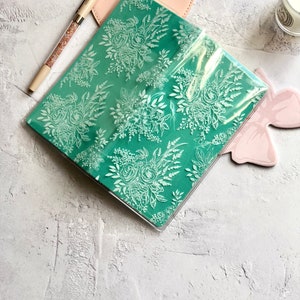 Puede incluir: Un cuaderno de color verde azulado con un patr&oacute;n floral blanco, cubierto con una funda de pl&aacute;stico transparente. El cuaderno est&aacute; sobre una superficie texturizada de color gris claro.