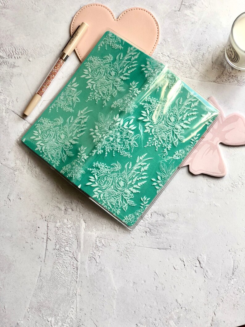 Puede incluir: Un cuaderno de color verde azulado con un patr&oacute;n floral blanco, cubierto con una funda de pl&aacute;stico transparente. El cuaderno est&aacute; sobre una superficie texturizada de color gris claro.