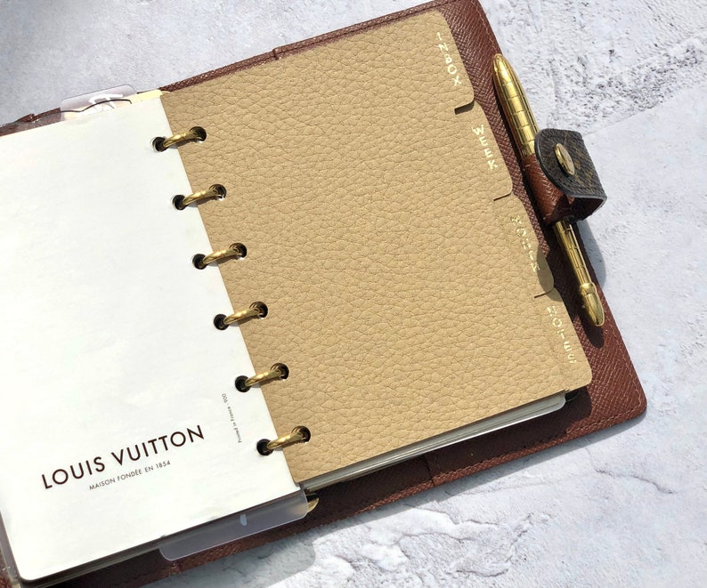 May include: A brown leather bound planner with gold rings and a beige insert. The insert has tabs labeled "Inbox", "Week", "Month", and "Notes". The planner has the Louis Vuitton logo and the text "Maison Fond&eacute;e en 1854".