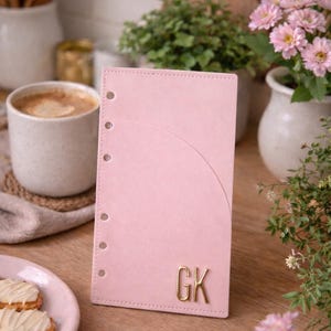 Könnte beinhalten: Ein hellrosa Lederplaner mit goldenen Initialen "GK". Der Planer hat eine gebogene Tasche und ist offen. Im Hintergrund eine Tasse Kaffee, Blumen und ein Teller mit Keksen.