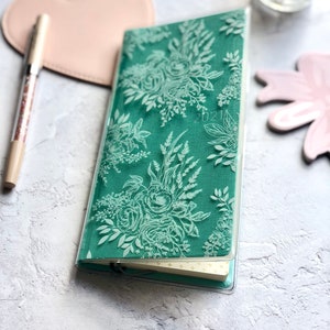 Puede incluir: Una cubierta de agenda de tela verde azulada con un dise&ntilde;o floral blanco. La cubierta es transparente y tiene la fecha 2021 impresa.