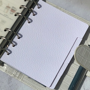 May include: A white, textured, blank page in a white leather bound planner with silver rings. A silver pen is lying on the planner.