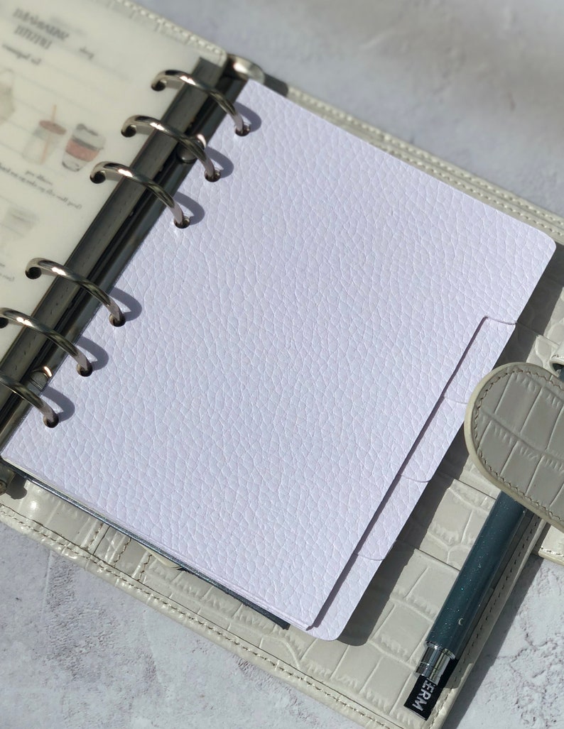 May include: A white, textured, blank page in a white leather bound planner with silver rings. A silver pen is lying on the planner.