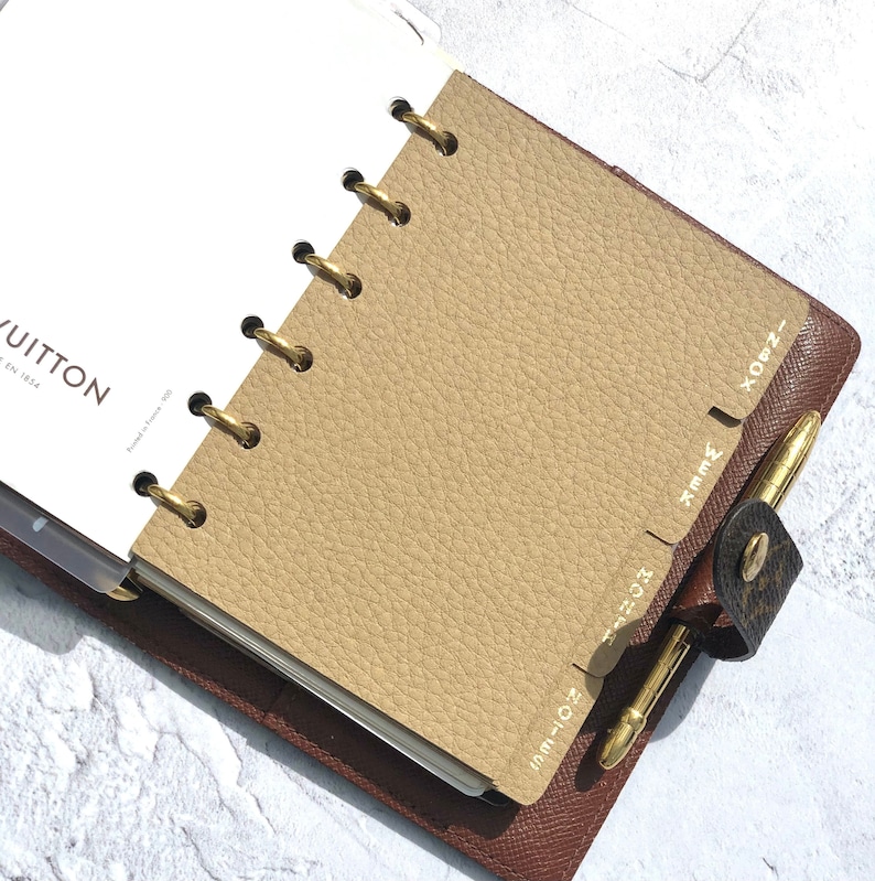 May include: A brown leather planner with gold rings and tabs labeled "OCT-NOV", "NOV-DEC", "JAN-FEB", "FEB-MAR", and "MAR-APR".