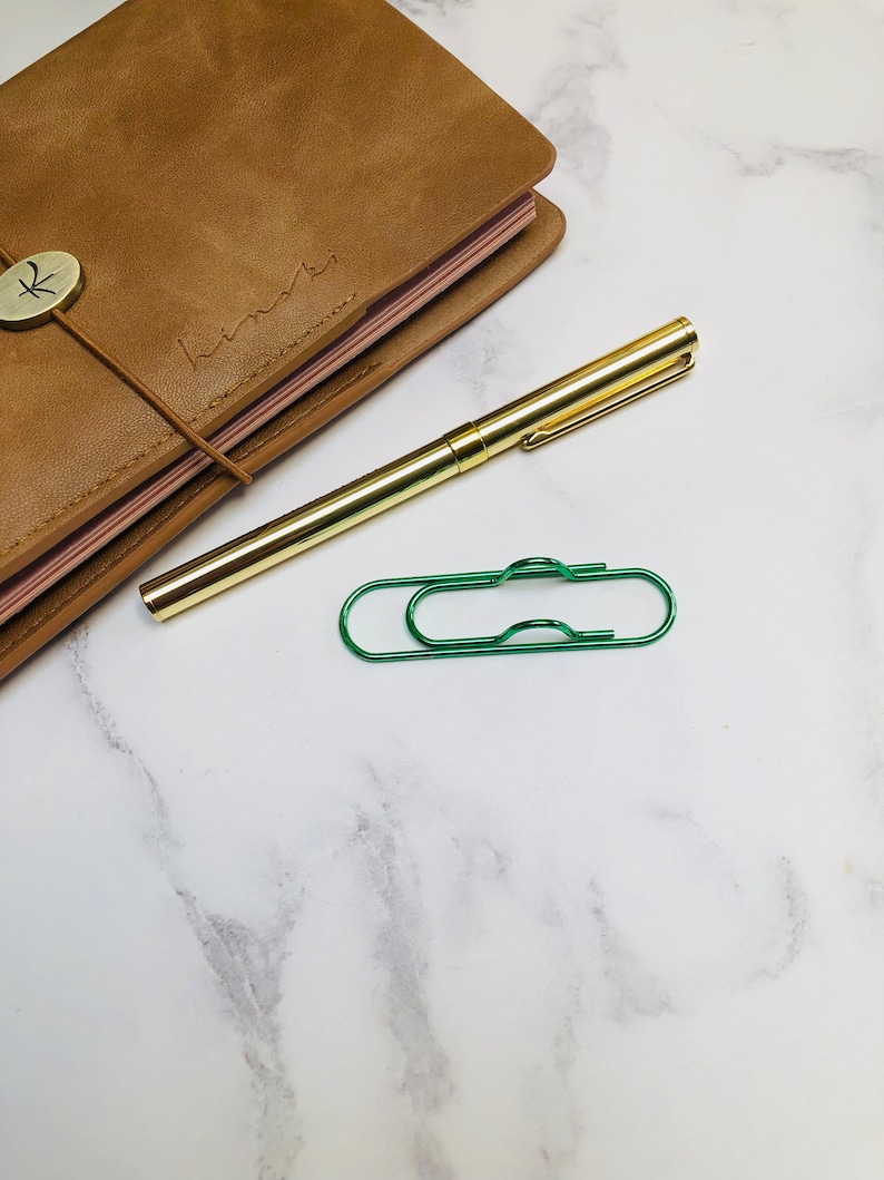 May include: A brown leather journal with a gold button and a gold pen on a white marble surface. A green paperclip is also on the surface.