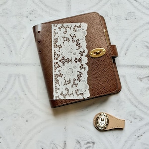 May include: A brown leather planner with a gold clasp and a white lace insert. A small beige leather bookmark with a floral design and pearl accents is also present. The planner is closed and has a textured surface.