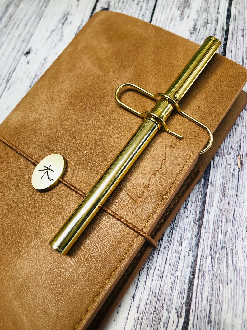 May include: A brown leather journal with a gold pen and a gold clasp. The journal has a small, round, gold tag with the Japanese character for "wood" on it. The journal is closed and the pen is tucked into the clasp.