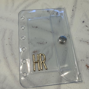 Puede incluir: Un tarjetero de plástico transparente con cierre de botón a presión e iniciales doradas "HR". El tarjetero tiene varios agujeros en el lado izquierdo para encuadernar. En el fondo se ven una cartera de cuero marrón y un bolígrafo dorado.