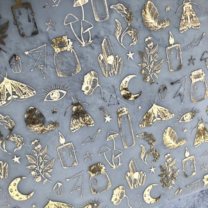 May include: A sheet of gold foil stickers featuring a variety of designs, including moths, mushrooms, crystals, candles, and stars. The stickers are on a light grey background.