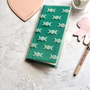 Op de afbeelding: Een turquoise planner met een transparante plastic hoes met een herhalend patroon van witte driedubbele maansimbolen. De planner is open op een pagina met het jaar 2021 erop gedrukt.