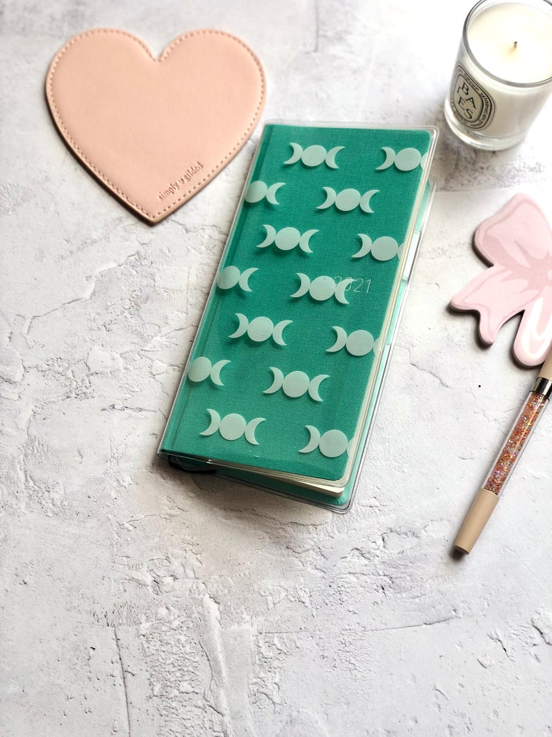 Op de afbeelding: Een turquoise planner met een transparante plastic hoes met een herhalend patroon van witte driedubbele maansimbolen. De planner is open op een pagina met het jaar 2021 erop gedrukt.