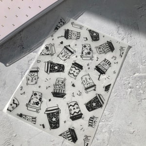 May include: White translucent sheet with a repeating pattern of black and white coffee cups. The cups have different designs, including stars, hearts, and musical notes.