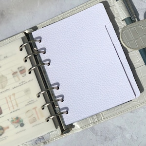 May include: A white, textured, blank page in a white leather bound planner with silver rings. The page has a tab on the right side.