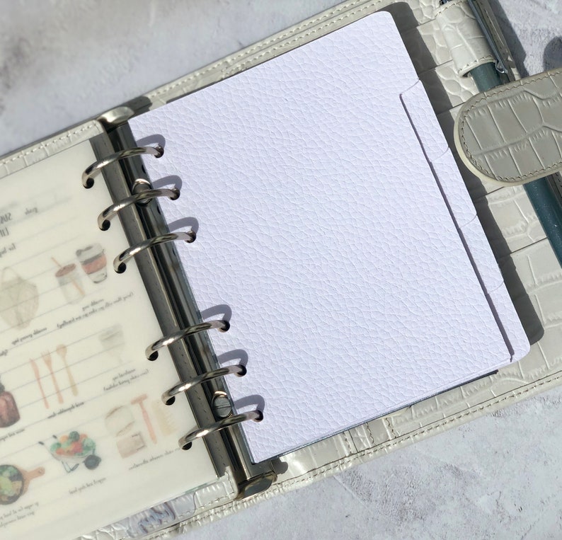 May include: A white, textured, blank page in a white leather bound planner with silver rings. The page has a tab on the right side.