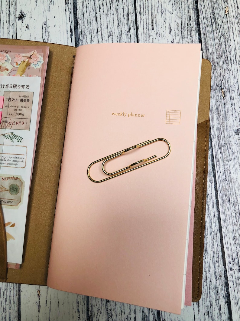 May include: A pink weekly planner with a gold paperclip on the cover. The planner has a brown leather cover and a white grid on the right side.