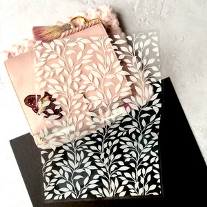 May include: A clear plastic sheet with a white leaf pattern. The sheet is lying on top of a pink leather notebook and a black surface.