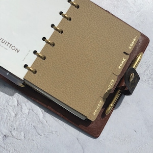 May include: A brown leather bound planner with gold rings and tabs. The tabs are labeled with the months of the year: Jun-Jul, Aug, Sep, Oct, Nov, Dec, Jan, Feb, Mar, Apr, May, and Jun. The planner has the Louis Vuitton logo on the front cover.