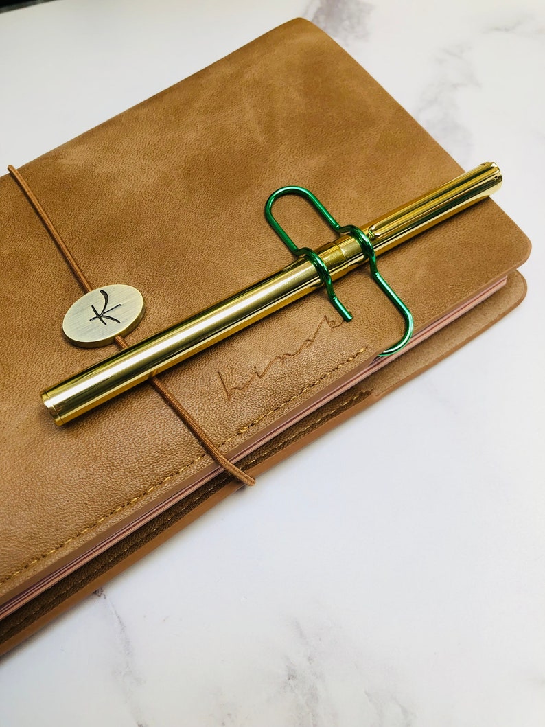 May include: A brown leather bound journal with a gold pen and a green metal clip. The journal has a gold metal button with a symbol on it. The journal is on a white marble surface.