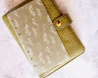 Puede incluir: Una agenda de imitación de cuero verde con una hoja de plástico transparente con un patrón floral blanco en el interior.