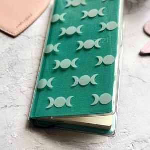 Op de afbeelding: Een turquoisegroen dagboek met een transparante plastic hoes. De hoes heeft een herhalend patroon van witte driedubbele maansimbolen. Het dagboek heeft het jaar 2021 op de voorkant gedrukt.