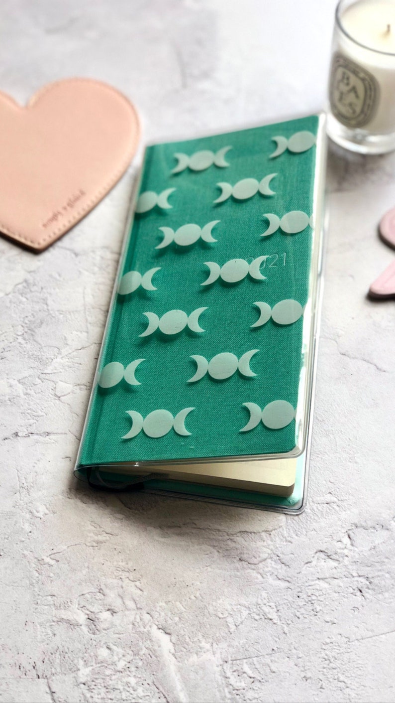 Op de afbeelding: Een turquoisegroen dagboek met een transparante plastic hoes. De hoes heeft een herhalend patroon van witte driedubbele maansimbolen. Het dagboek heeft het jaar 2021 op de voorkant gedrukt.