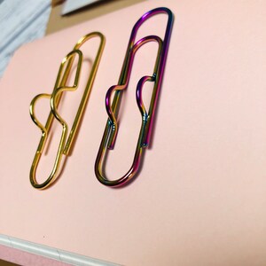 May include: Two paper clips, one gold and one iridescent, are on a pink surface. The iridescent paper clip has a rainbow finish.