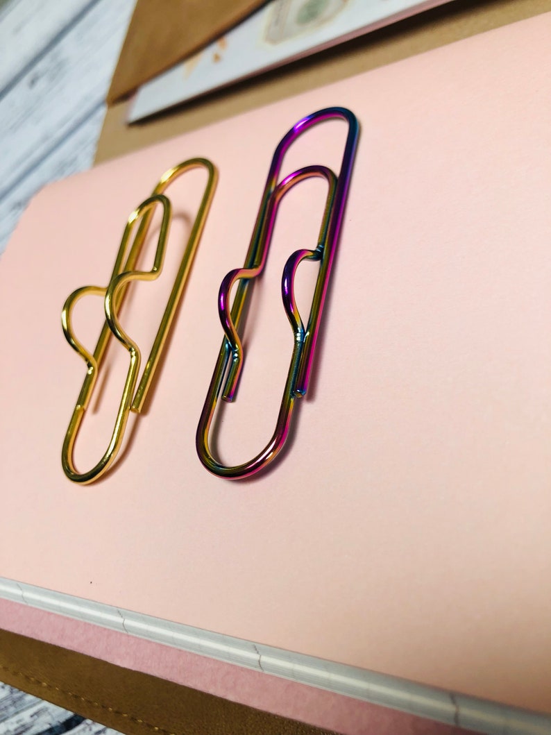 May include: Two paper clips, one gold and one iridescent, are on a pink surface. The iridescent paper clip has a rainbow finish.