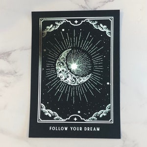 May include: A black card with a silver crescent moon and starburst design. The text "FOLLOW YOUR DREAM" is printed at the bottom of the card.