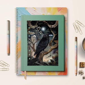 May include: A notebook featuring a crow illustration, alongside a pen, pencil, paper clips, and a coffee cup. The notebook has a green cover with a crow perched on a branch under a full moon. The pen and pencil have a rainbow gradient.