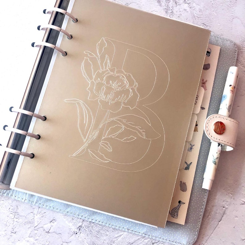 May include: A gold planner with a white floral design and the letter "B" in the center. The planner has a ring binding system and a white pen is attached to the side.