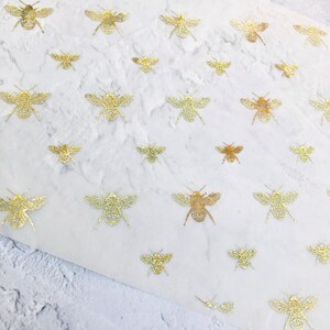 Puede incluir: Una hoja de papel transparente con un patrón repetitivo de abejas de lámina dorada. Las abejas están todas orientadas en la misma dirección y están dispuestas en un patrón escalonado.