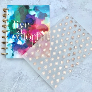 May include: A clear plastic sheet with a repeating pattern of gold dandelion designs. The sheet is laying on top of a colorful planner with the words "live a colorful life" printed on the cover.