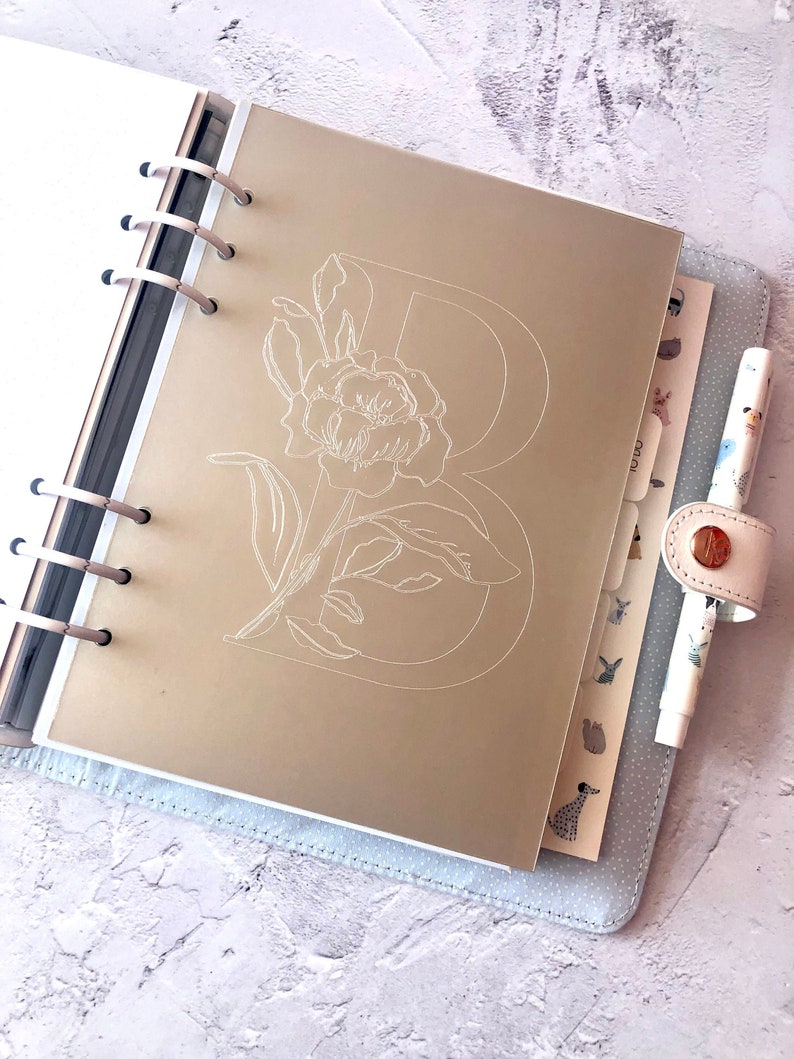 May include: A gold planner with a white floral design and the letter "B" on the cover. The planner is open to a page with a white floral design.
