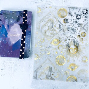 Op de afbeelding: Een kleine planner met een paars en blauw aquarelontwerp en de woorden "Born to Wander". Het heeft een zwarte elastische band met witte stippen en een zwarte kattenbedel. Daarnaast een transparante hoes met gouden hemelse ontwerpen.