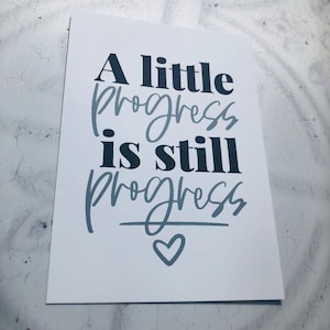 May include: A white card with the text "A little progress is still progress" in black and grey lettering. A small grey heart is drawn below the text.