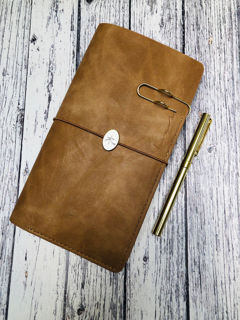 May include: A brown leather journal with a metal clasp and a small metal tag with a design on it. A gold pen is lying next to the journal.