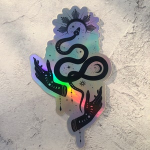 May include: A holographic sticker featuring a black silhouette of two hands holding a snake that is curled into an infinity symbol. The snake is surrounded by stars and a crescent moon. Above the snake is a sun with rays.
